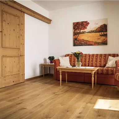 A cozy living room with a red sofa and a wooden table. A landscape painting hangs on the wall, and the room has plenty of natural light.