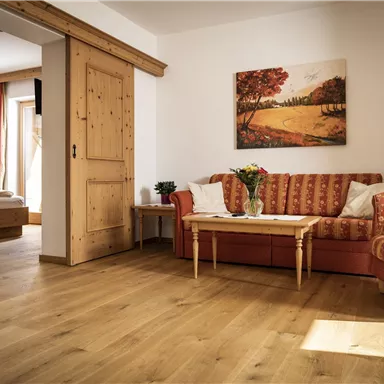 A cozy living room with a red sofa and a small table. A painting of a landscape hangs on the wall, and the wooden floor creates a warm atmosphere.
