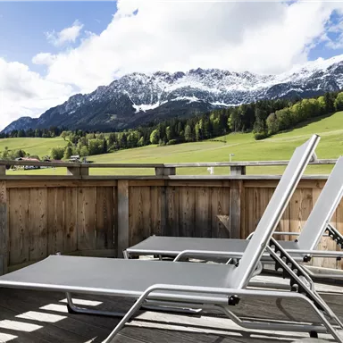 A balcony with lounge chairs and a view of green meadows and snow-capped mountains. The sky is clear and sunny.