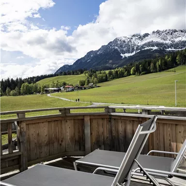 A balcony with two loungers offers a view of green meadows and majestic mountains. The sky is partly cloudy, and the scene exudes tranquility.
