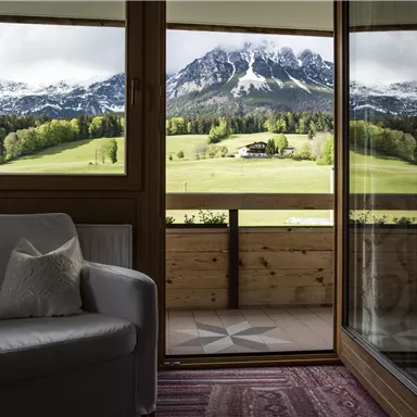 A cozy room with a white armchair and a view of a green landscape with mountains in the background. The view shows clouds over snow-capped peaks.