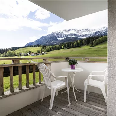 A cozy balcony with two white chairs and a table. In the background, green meadows and majestic snowy mountains can be seen.