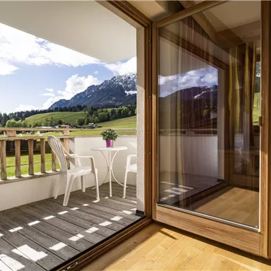 A cozy balcony with two white chairs and a table. In the background, green meadows and mountains can be seen under a clear sky.