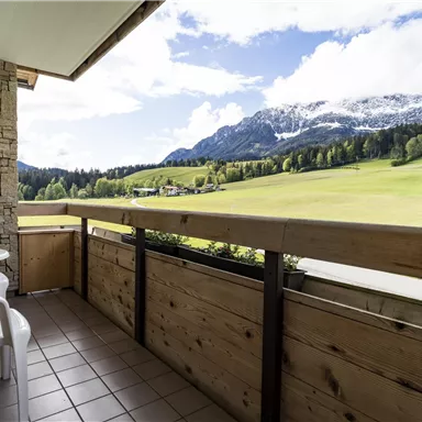 A balcony with two chairs and a view of green meadows and snow-capped mountains. The sky is partly cloudy and the landscape is picturesque.