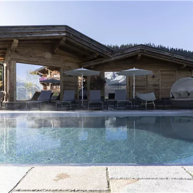 A modern pool area with steaming water and a wooden terrace. In the background, there is a stylish wooden house and some loungers.