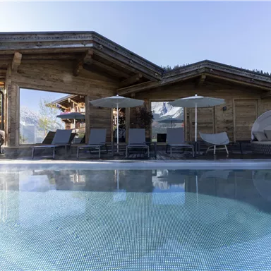 A modern wooden house with a terrace and loungers. In the foreground is a pool that reflects the landscape.