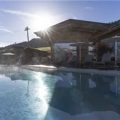 A beautiful outdoor pool with sun umbrellas and loungers, surrounded by a picturesque landscape. The sun shines over the huts in the background.