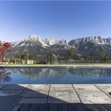 A beautiful pool with a view of majestic mountains and a clear blue sky. The surroundings are green and inviting, perfect for relaxing.