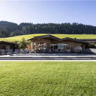 A modern alpine hut with a spacious garden and pool. In the background, green hills and trees can be seen.