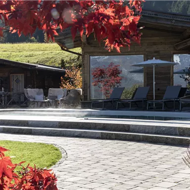 A beautiful outdoor area with loungers and sun umbrellas surrounded by colorful foliage. In the background, there are wooden-clad buildings and a green meadow.