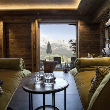 A cozy room with modern sofas and a round table. The large window front offers a breathtaking view of the mountains.
