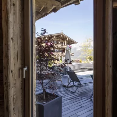 A view through a window onto a terrace with loungers. In the background, mountains and a blue sky are visible.