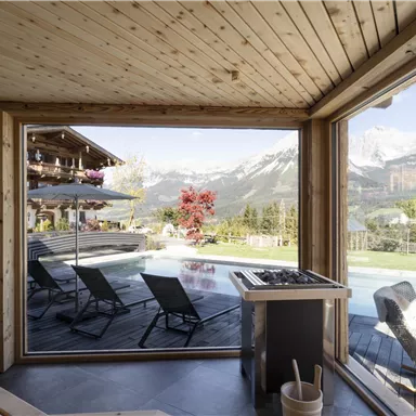 A modern holiday home with wooden elements and large windows. You can see the outdoor pool and the surrounding mountains in the background.