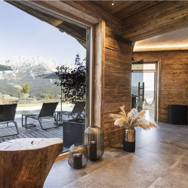 A modern interior with wooden elements and large windows that offer a view of the mountains. Outside, deck chairs and sun umbrellas can be seen.