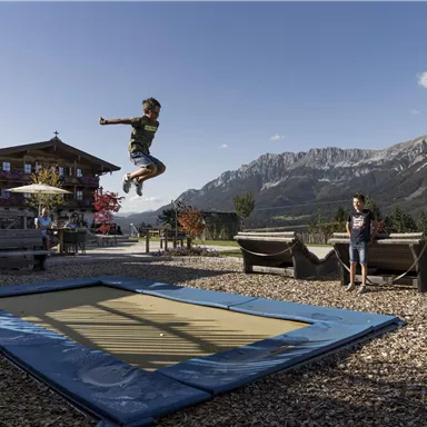 A boy jumps high on a trampoline, surrounded by a beautiful mountain panorama. In the background, there is a mountain hut and seating options.