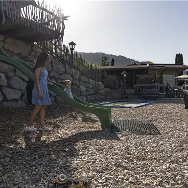 A playground with a green slide and pebble ground. In the background, there are seating areas and a shady spot visible.