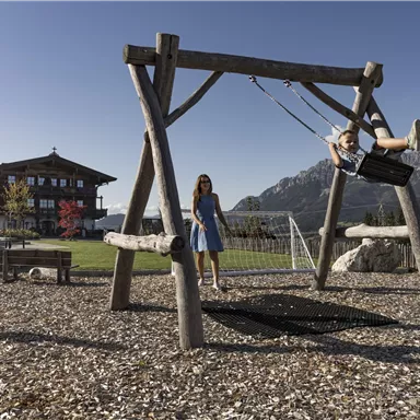 A playground with a swing and two children. In the background, mountains and a beautiful wooden house can be seen.