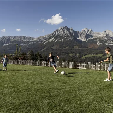 Three children are playing soccer on a green lawn with impressive mountains in the background. The sky is clear and the landscape is picturesque.