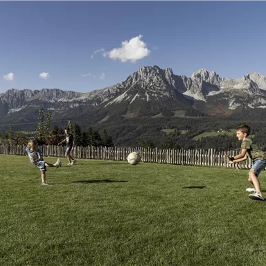 Children are playing soccer on a green meadow with mountains in the background. The sky is clear and blue, creating a relaxed atmosphere.