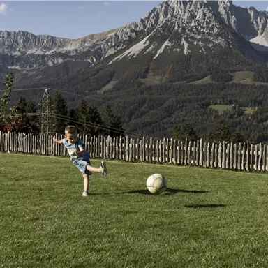 A boy is playing with a soccer ball on a green meadow. In the background, impressive mountains and a clear sky can be seen.
