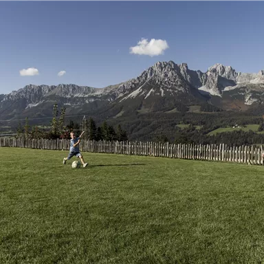 Two children are playing soccer on a green meadow with mountains in the background. The sky is clear and sunny.