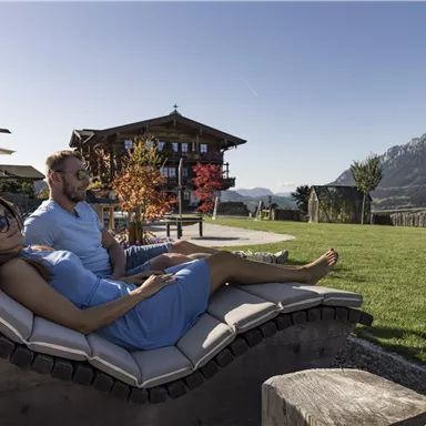 A couple relaxes on loungers in a beautiful garden with a view of the mountains. In the background, a traditional chalet and a clear sky can be seen.