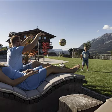 A family is playing in the garden while the sun is shining. In the background, mountains and a beautiful house can be seen.