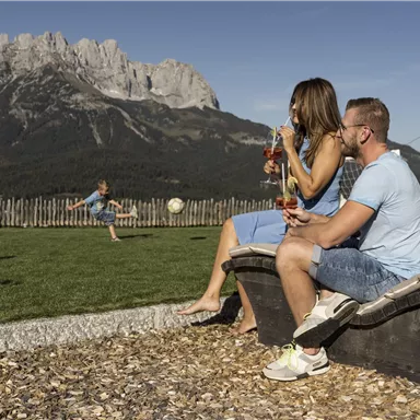 A couple sits relaxed in a hammock and enjoys drinks. In the background, a child plays soccer in front of an impressive mountain landscape.