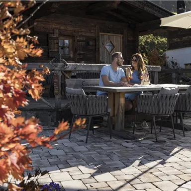 A cozy terrace with a table and chairs, surrounded by colorful autumn leaves. A couple sits together and enjoys drinks in the sunshine.
