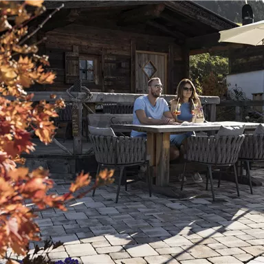 A couple sits relaxed at a table outdoors in front of a rustic wooden cabin. The surroundings are autumnal with colorful leaves and sunny weather.