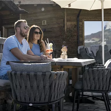 A couple is sitting relaxed at a table outdoors. In the background, cozy wooden houses and sun umbrellas can be seen.