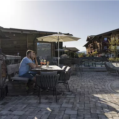 A couple is sitting at an outdoor table enjoying drinks. In the background, alpine-style buildings and a sunny landscape are visible.