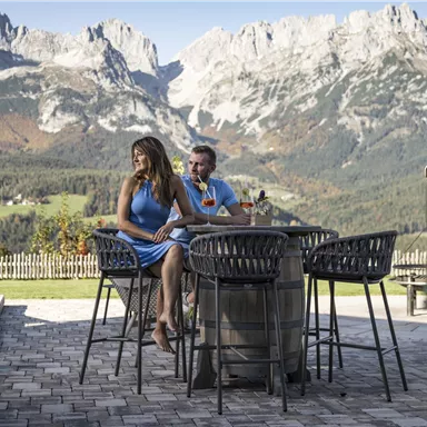 A couple sits at a table outdoors with a view of impressive mountains. The scenery is surrounded by beautiful landscape and radiates a relaxed atmosphere.