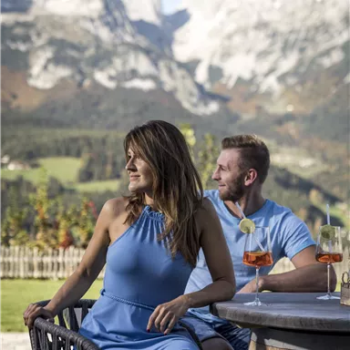 A couple sits at a table with drinks, enjoying the view of the mountains. In the background, green meadows and snow-capped peaks can be seen.
