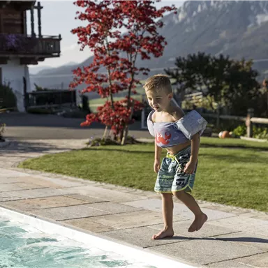 A small boy with swim wings stands at the edge of a pool. In the background, a beautiful house and trees can be seen.