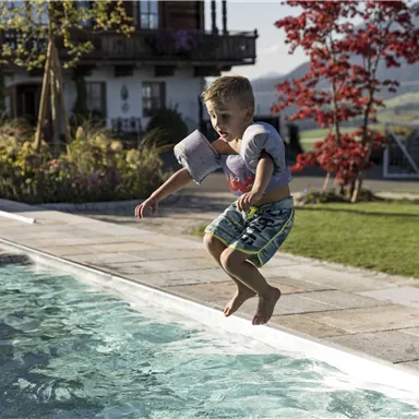 A boy jumps happily into a clear pool. In the background, green meadows and beautiful plants can be seen.