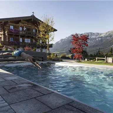 A man jumps into a swimming pool in front of a beautiful alpine landscape. In the background, a traditional wooden house and colorful trees are visible.