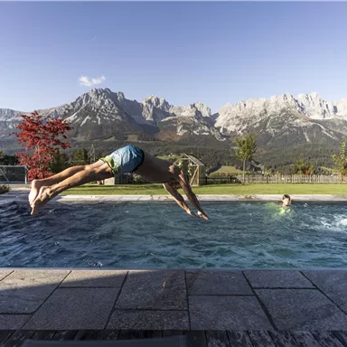 A man jumps into a swimming pool against an impressive mountain backdrop. In the pool swims another person, surrounded by trees and a clear landscape.