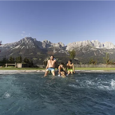 A family is splashing in the water of a pool with a view of majestic mountains. The sun is shining and the sky is clear.