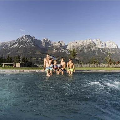 A family is sitting at the edge of a swimming pool with a view of majestic mountains. It is a sunny day and the surroundings are green and inviting.