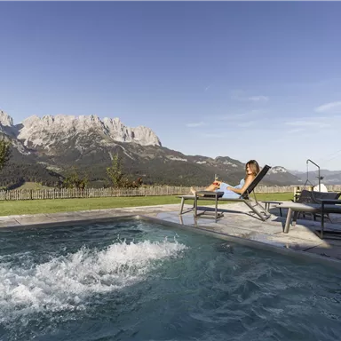 A beautiful pool with bubbling water and a view of the mountains. A person is relaxing on a sun lounger in the sun.