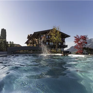 A pool in front of a charming wooden house with a clear blue sky. In the background, mountains and colorful trees can be seen.