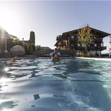 A beautiful outdoor pool with two children playing in it. In the background, there is a charming building and a clear sky visible.