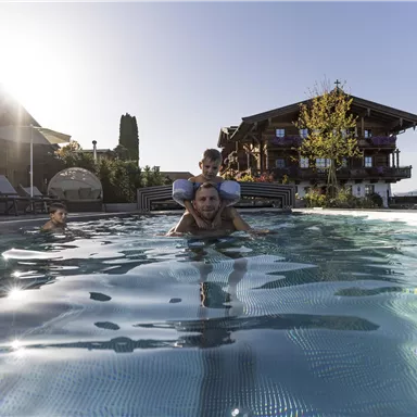A pool with several people having fun in the water. In the background, there is a cozy hotel and beautiful plants visible.