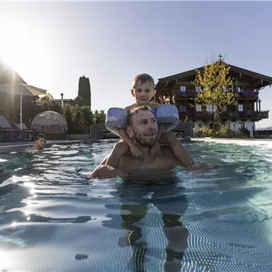A father is relaxedly swimming in the pool with his son on his shoulders. In the background, a cozy wooden house and colorful trees can be seen.