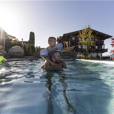 A man is swimming in the pool with a child on his shoulders. In the background, a beautiful house and the mountains can be seen.