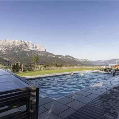 A pool with a view of impressive mountains and a green landscape. The sky is clear and the sun is shining.