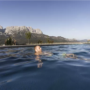 A relaxing pool with a view of impressive mountains. A person is swimming relaxed in the water under a clear sky.