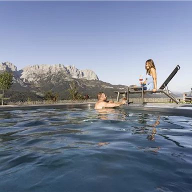 A man and a woman are enjoying a relaxing moment by the pool with the mountains in the background. The sky is clear and the atmosphere is calm and inviting.