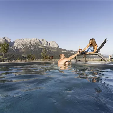A man and a woman are enjoying a relaxed moment by the pool. In the background, majestic mountains and a clear sky can be seen.
